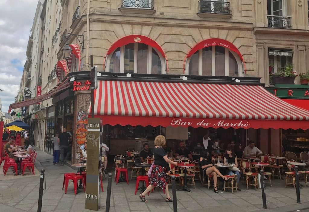 Bar du Marché, a brick bar with red awning, tables, and chairs on the corner of the rue de Seine and rue de Buci. The view is from the street and there are people enjoying a drink at tables outside