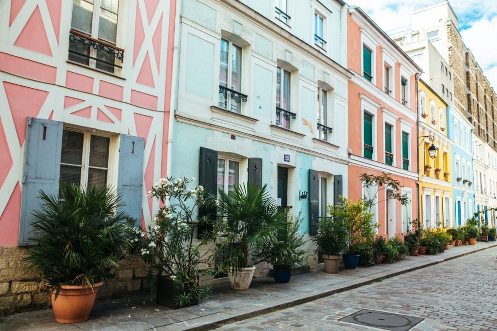 An angled view of a street of pastel houses, one is pink with white geometrical designs, the next robin egg blue, then orange, yellow, and again blue