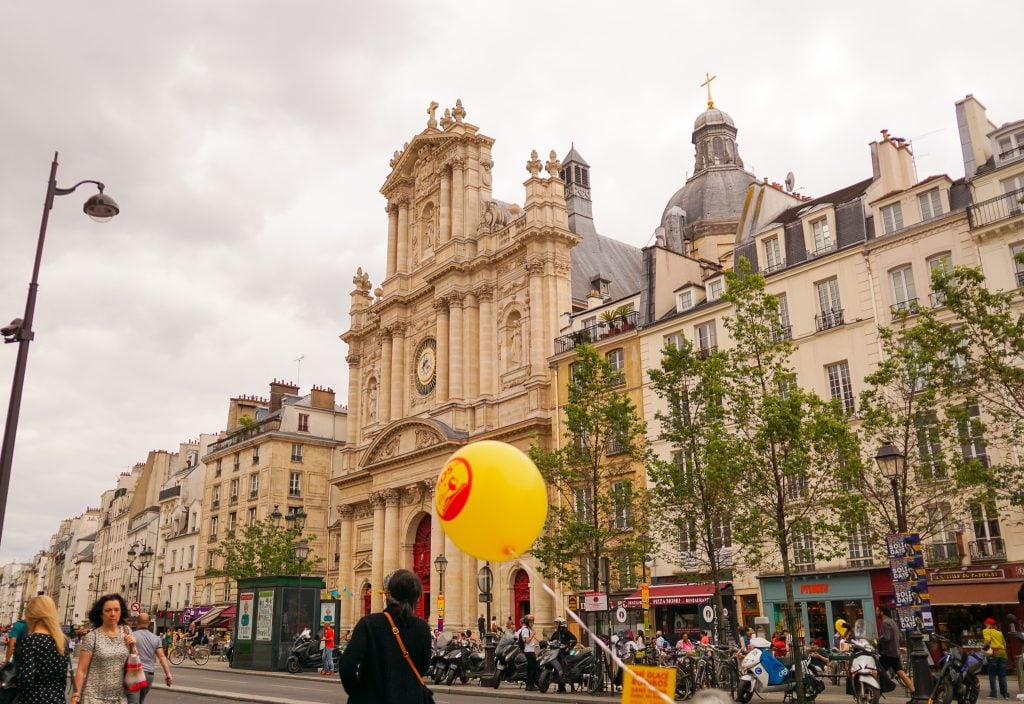 A view of Rue Saint Antoine on a cloudy day. A child is carrying a bright yellow balloon up the street and there are trees growing in front of a building.