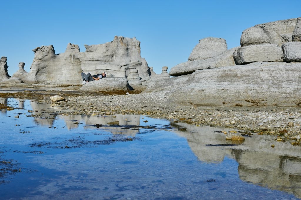 Gray rock formations popping up from a beach underneath a blue sky. A person relaxes, lying on one of the rocks.