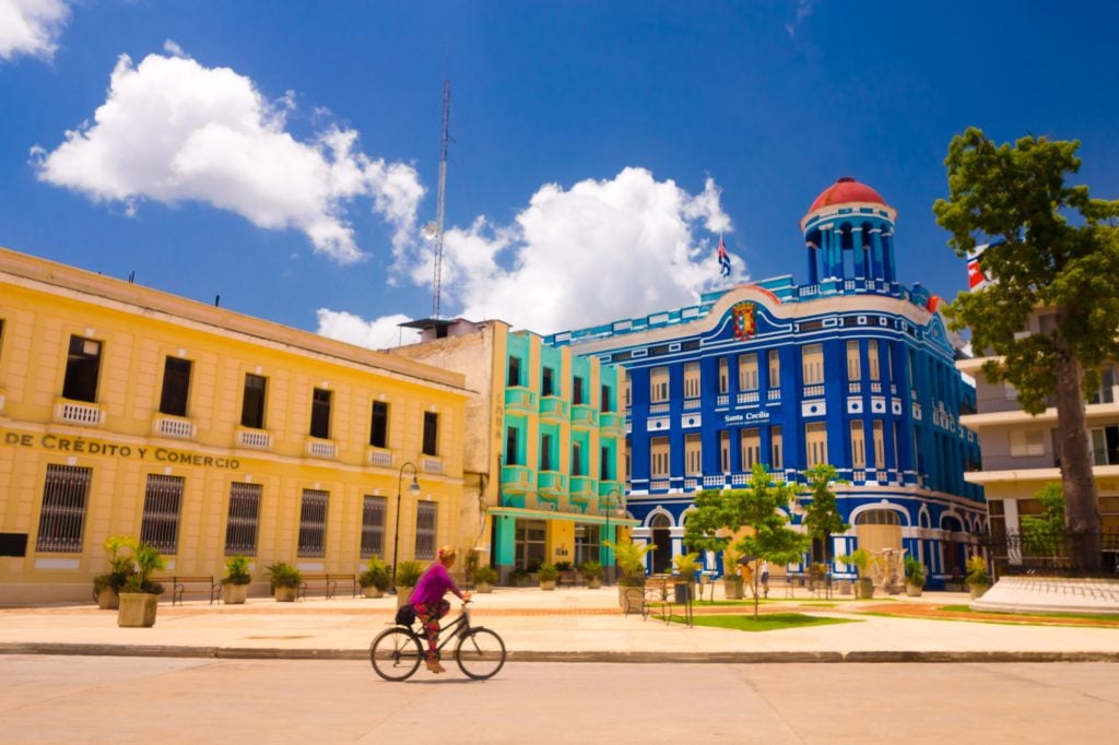 A woman cycling down a plaza with several colonial buildings, including one bright green and one deep blue.