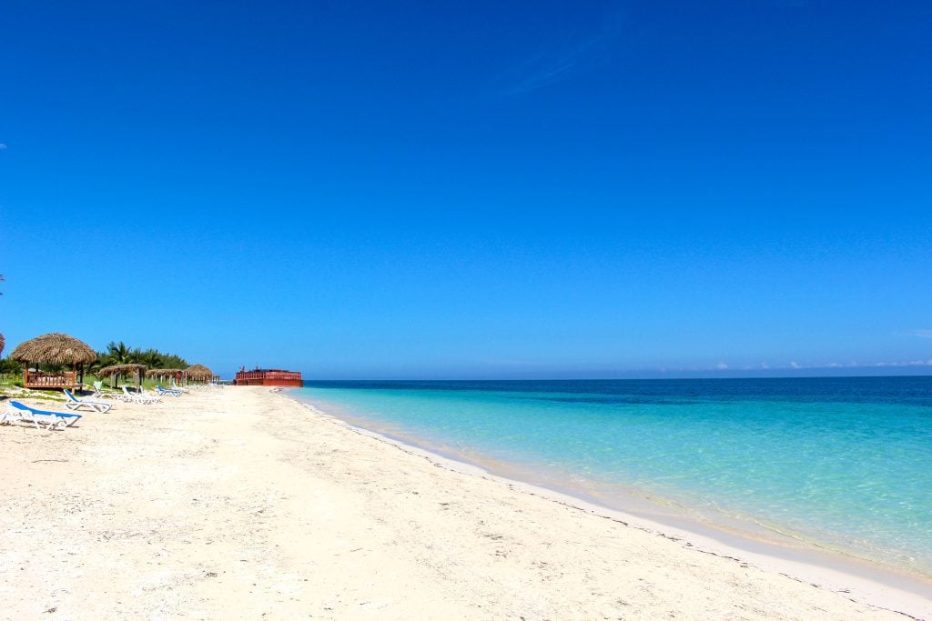 A strip of untouched beach, white sand next to bright blue water, a few beach chairs and palaces on the beach.