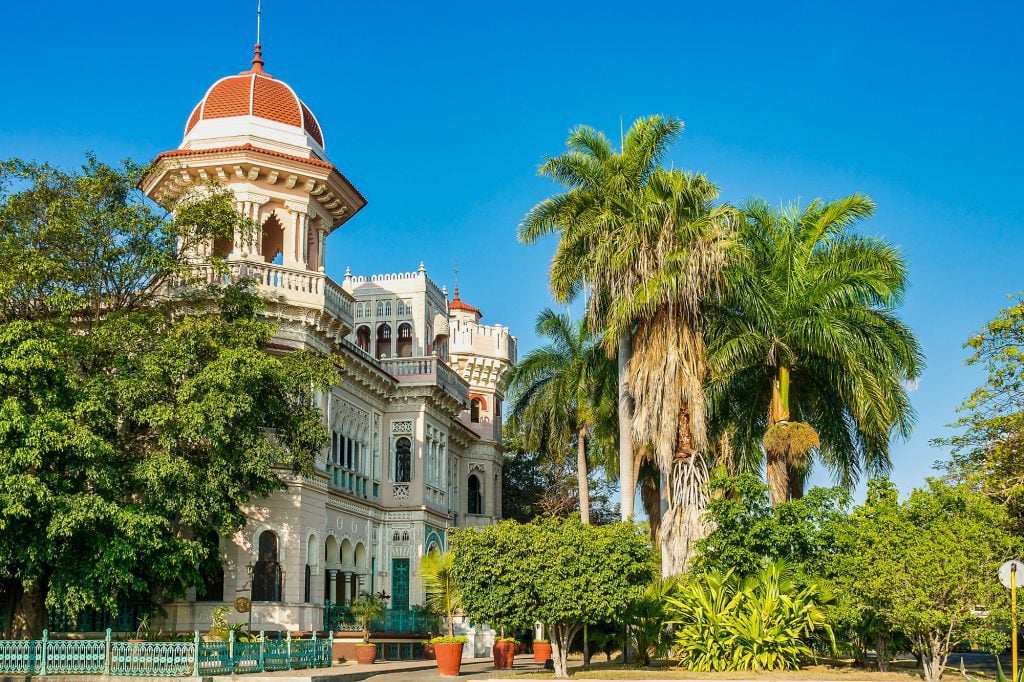 A white colonial building with lots of columns and zig-zag lined windows among several palm trees.