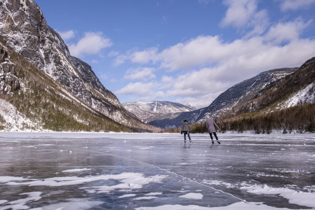 Two ice skaters skate across a frozen river between snow-dusted mountains, underneath a partly cloudy sky.