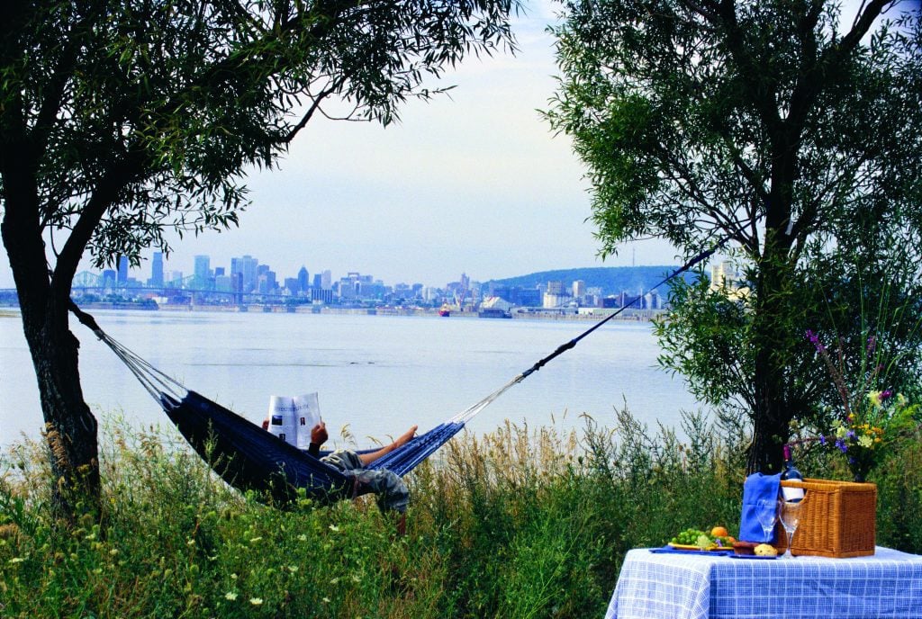 A person in a hammock strung between two trees, reading a newspaper, as you can see the river and the Montreal skyline in the distance.