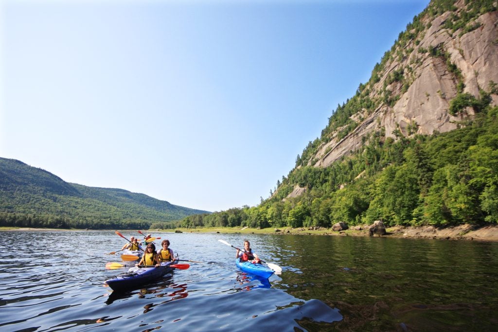 Three kayaks paddling down a river as a mountainous fjord rises up on their right.