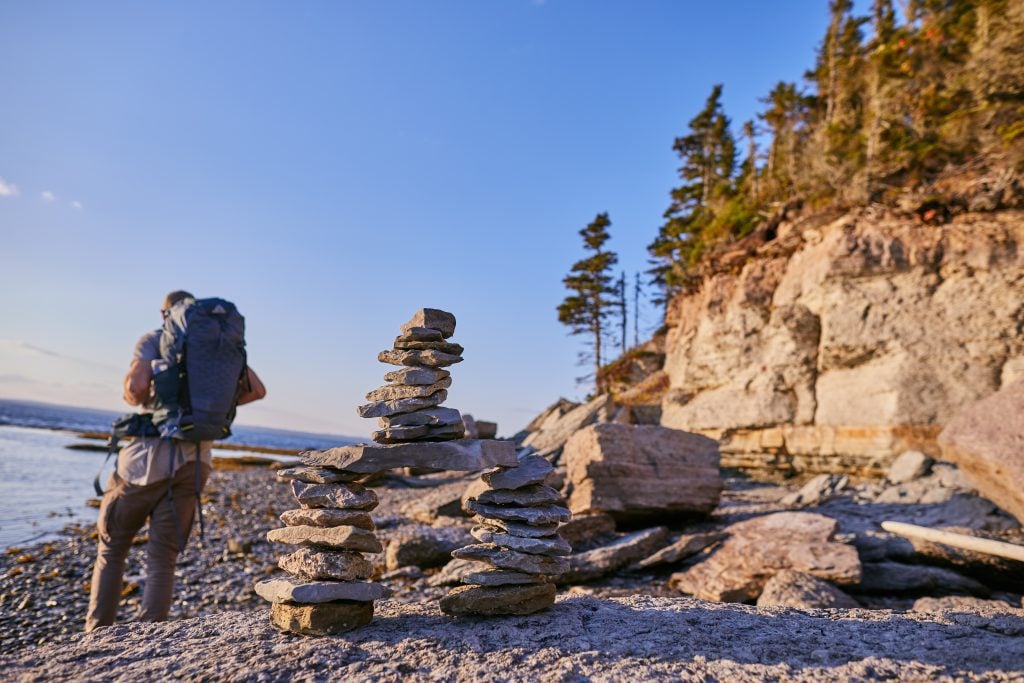 A lone hiker standing on a beach with a backpack. In front of him is a manmade rock cairn. You can see tall pine trees and steep limestone cliffs, and still water in the distance.