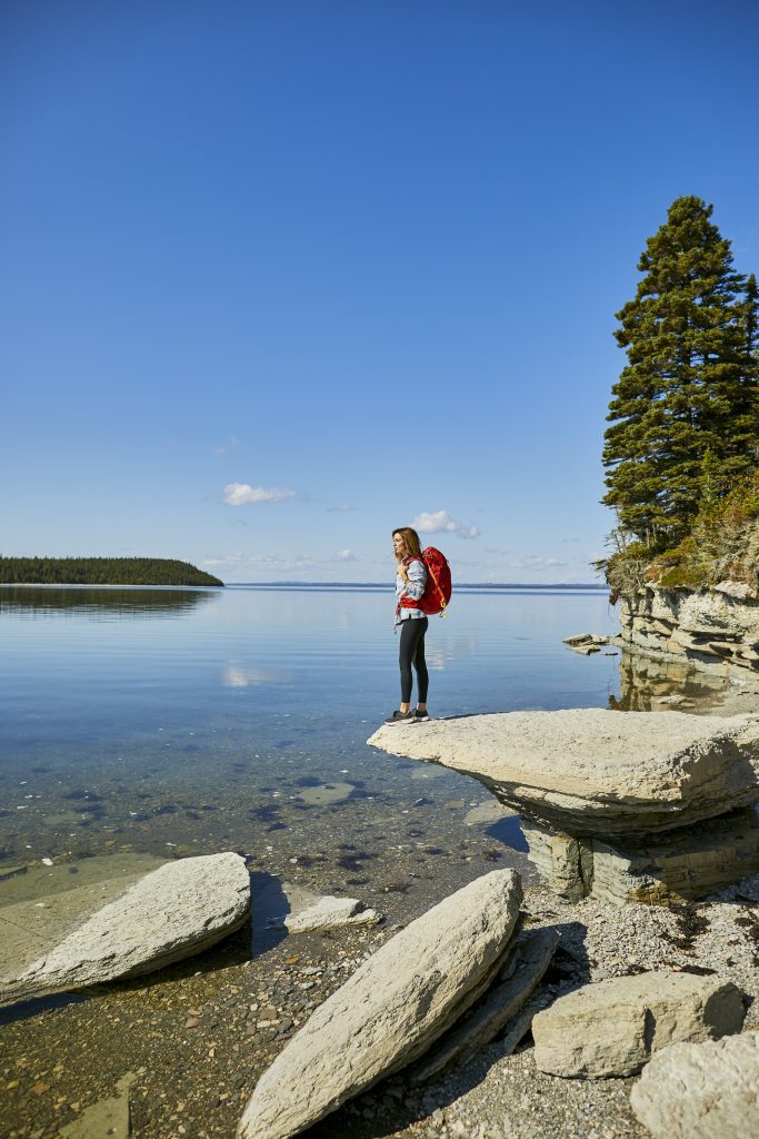 A woman hiker stands on a jutting out flat pancake rock over a still sea with clear water, forested land in the distance.