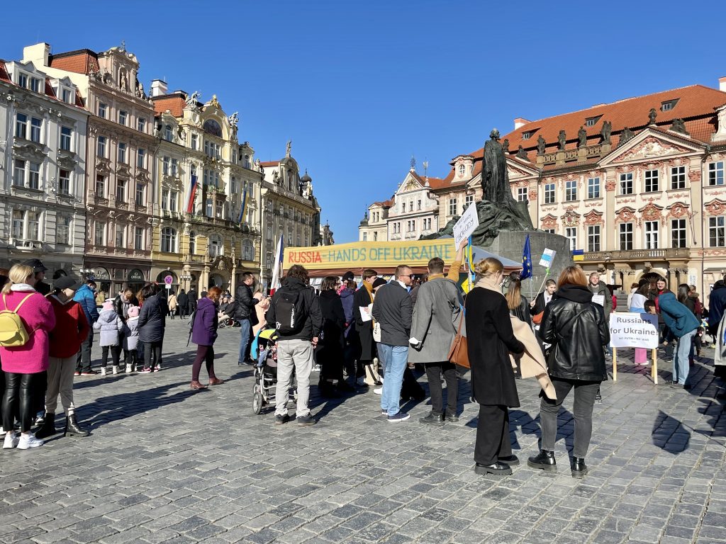 The pastel-colored crenellated buildings of Old Town Square in Prague. In the middle is a sign reading "Russia -- Hands Off Ukraine" and a crowd around it. One sign reads "Russians for Ukraine."