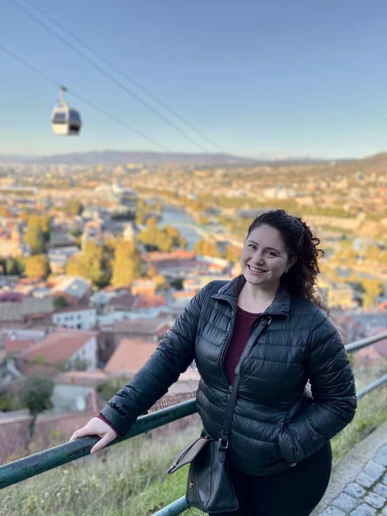 Richelle standing in Tbilisi, Georgia, a cable car in the air behind her, and a blurry city view over orange roofs and the river.