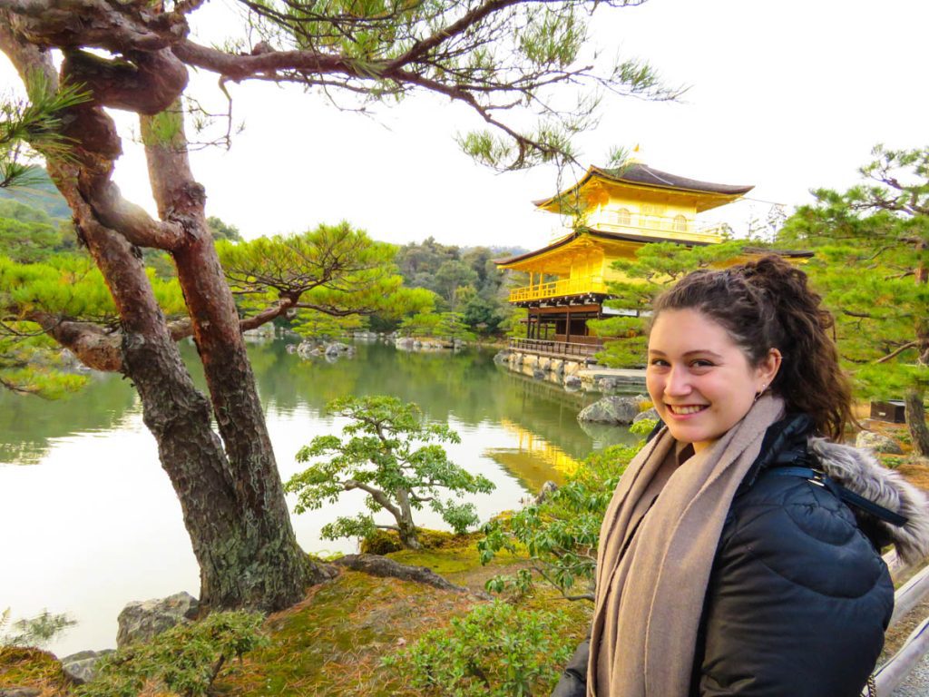 Richelle, smiling with dark curly hair, a black coat, and long scarf, standing in front of the bright gold temple in front of a lake in Kyoto, Japan.