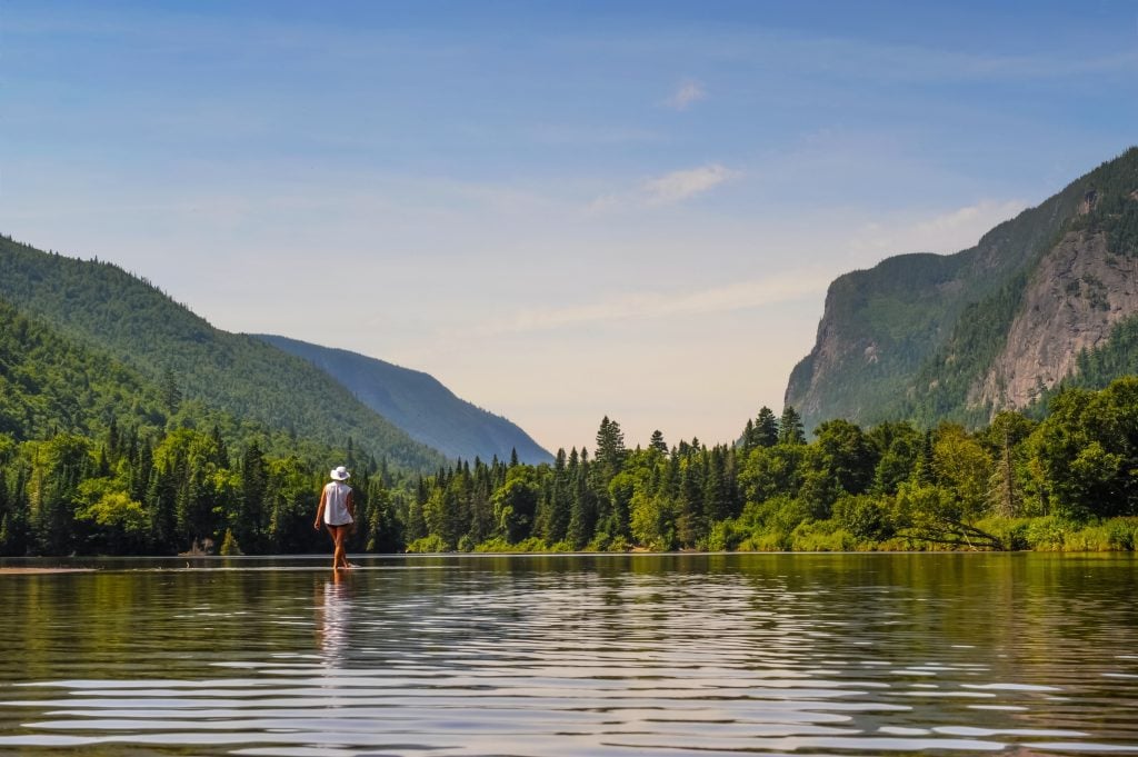 A person stands in shallow, still water (looking like she's walking on water), trees and rock formations in the distance.