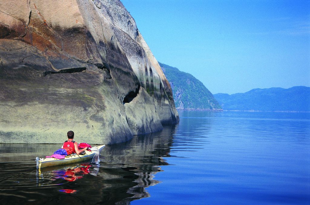 A lone kayaker paddling along still blue water as rock formations rise up around him.