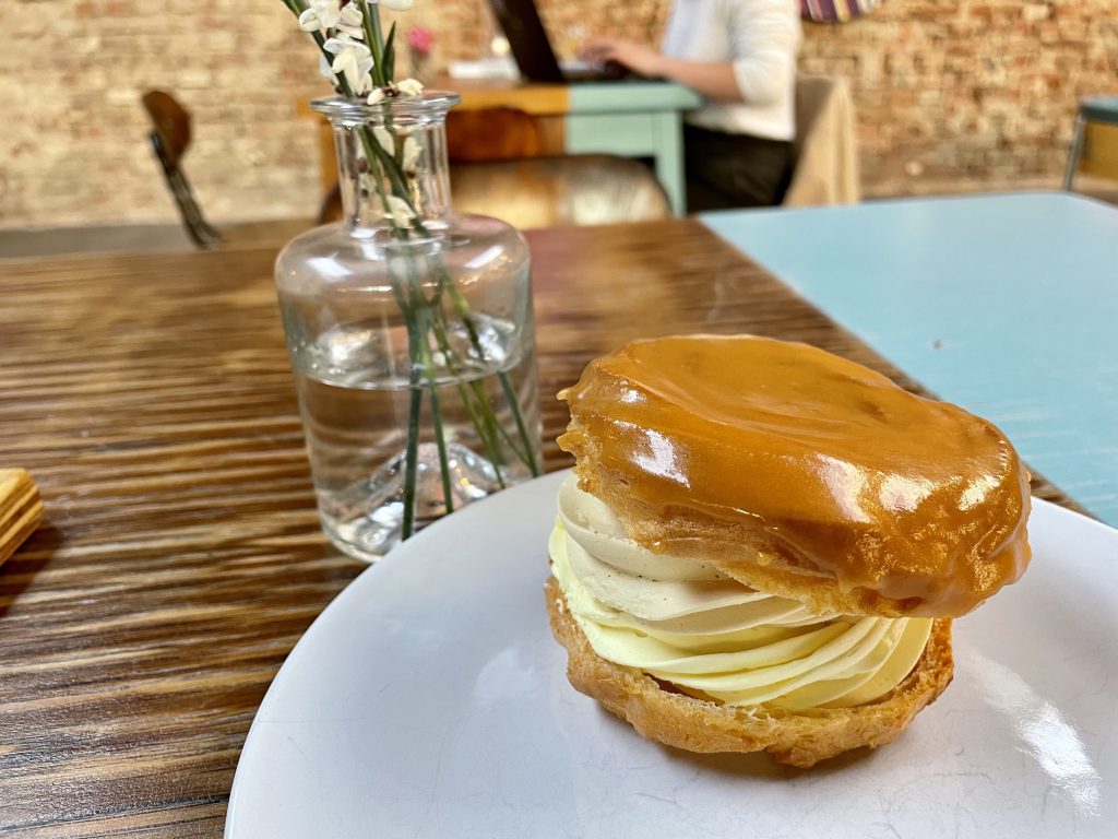 A pastry on a plate: what looks like a cream puff with two kinds of swirled pastry cream inside (caramel and regular), topped with a caramel glaze. It's on a plate next to a small vase of flowers.