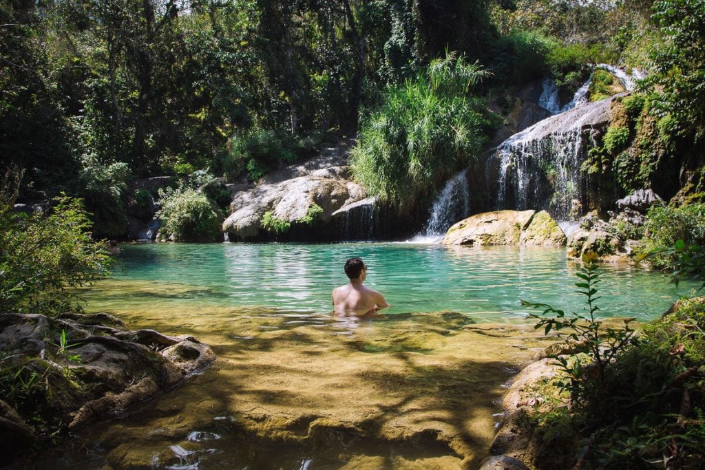 A person swimming in clear blue-green water with waterfalls running into it