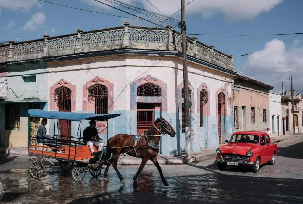 A horse drawn carriage and an antique red car on the corner of a Cuban street