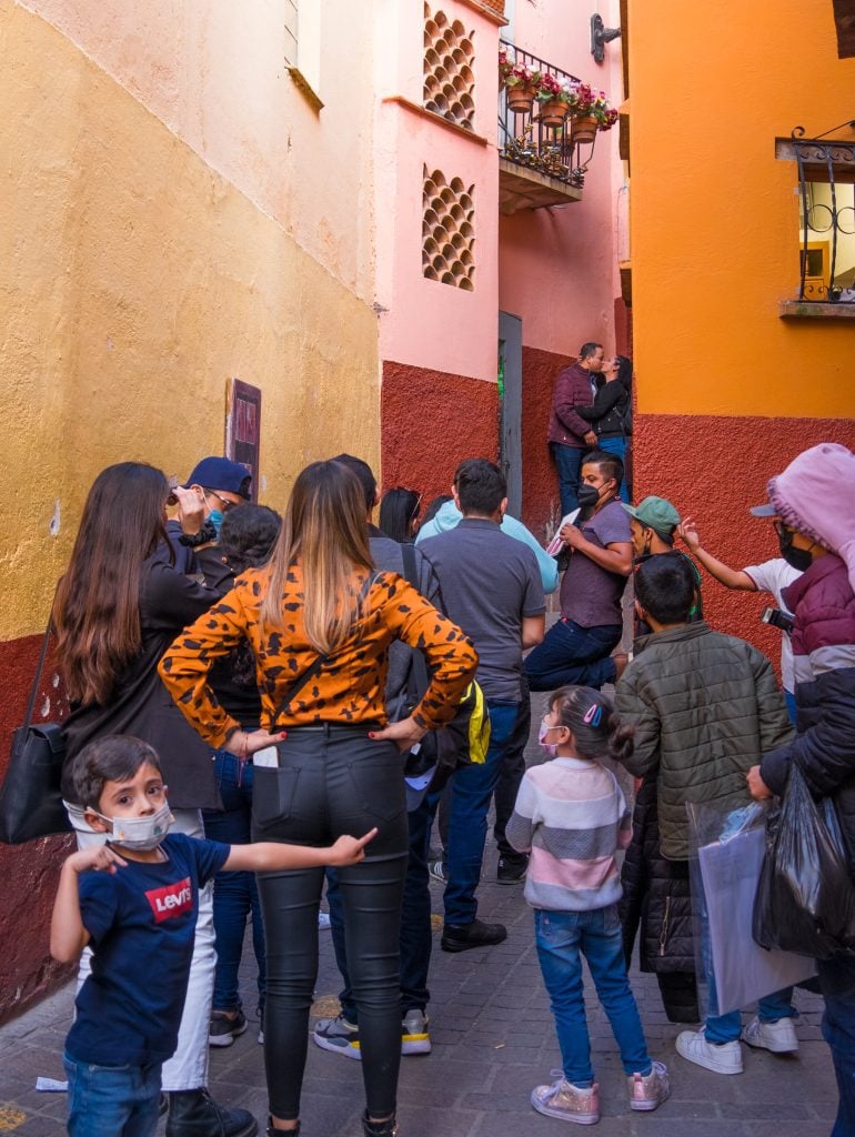 A couple kissing in the alley of the kiss, a big crowd lined up behind them. In front a little boy in a face masks points at them as if to say, Look!