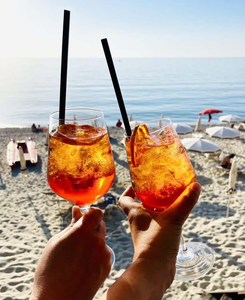 Two bright orange aperol spritzes being toasted in front of a beach.