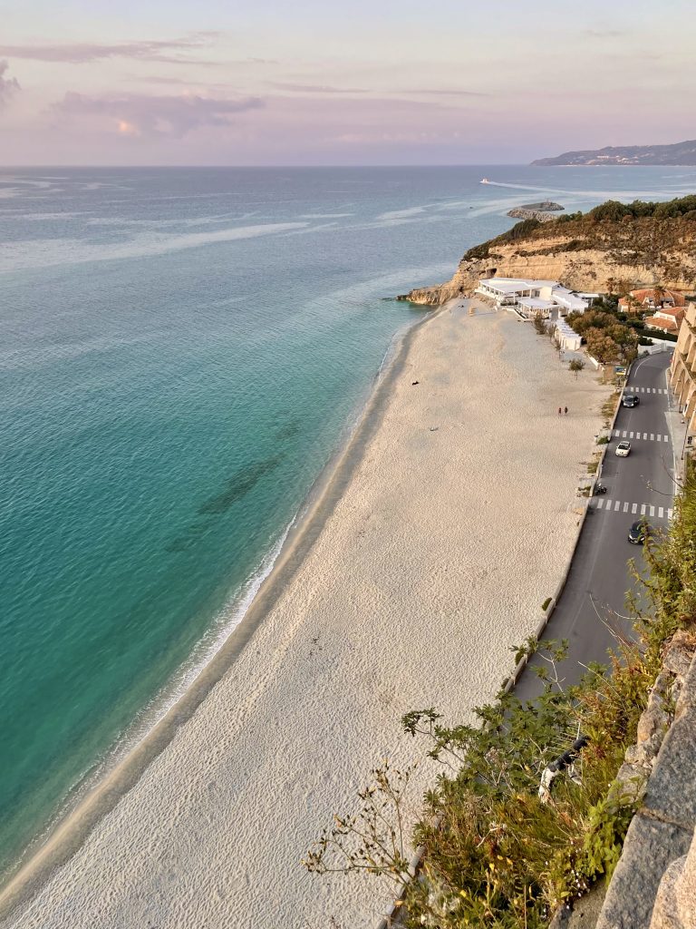 A long, nearly empty sandy beach in Tropea, next to clear turquoise water with gentle waves.