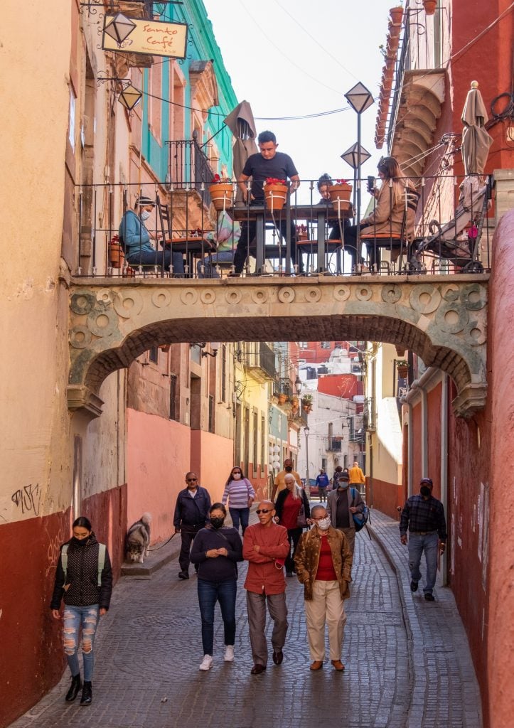 A bridge between two buildings that also serves as an outdoor restaurant. A waiter serves people sitting at tables. Below, people walk down the street, most of them masked.