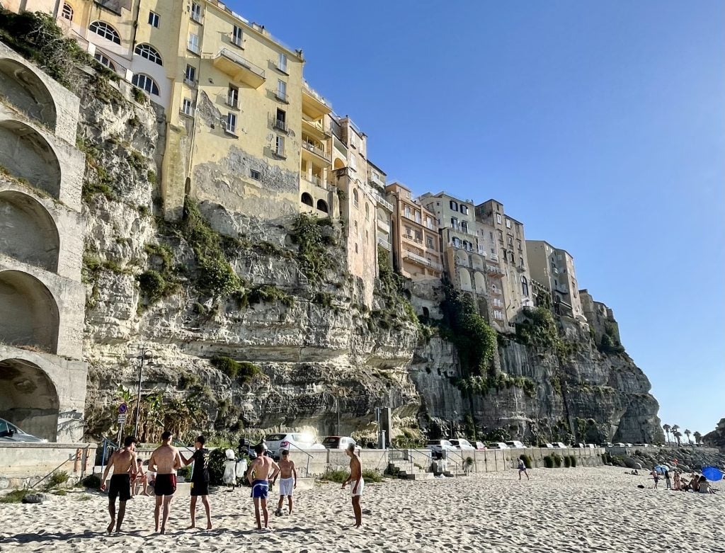 A group of shirtless teenage boys playing soccer on the beach, the cliffs of Tropea behind them.