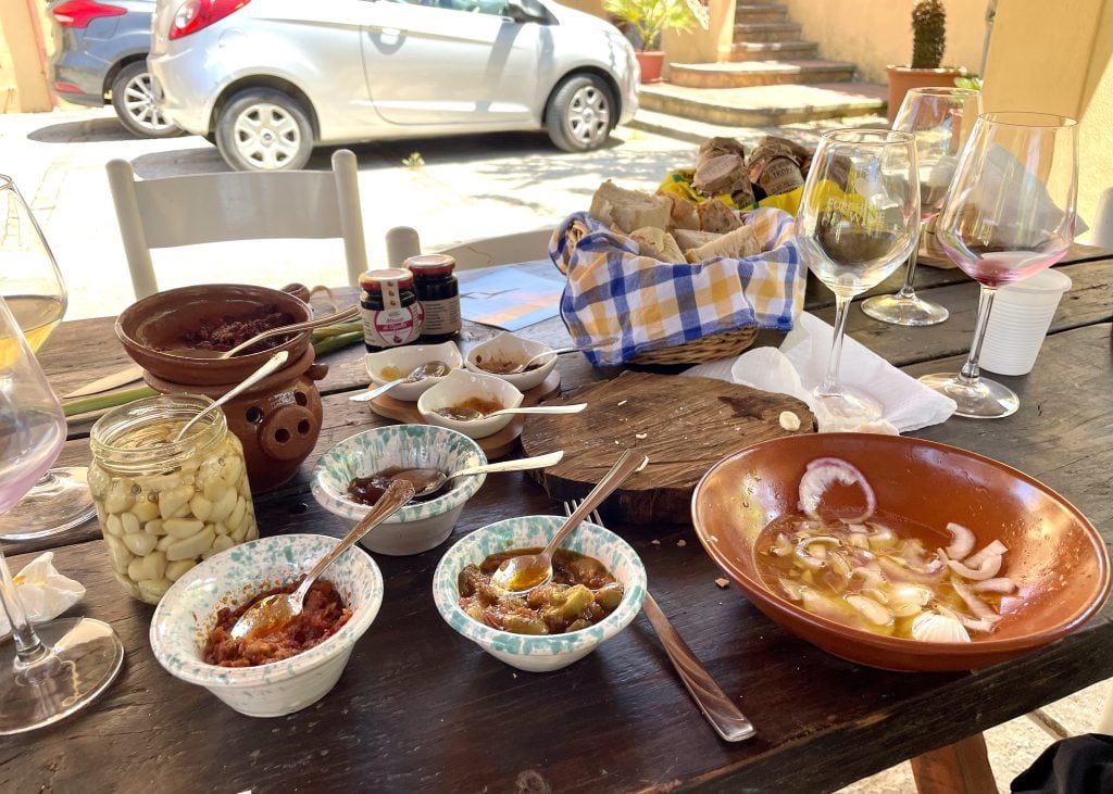 Several tasting plates filled with onion salad, lots of jams and spreads, pickled garlic, wine glasses, and a basket of bread.