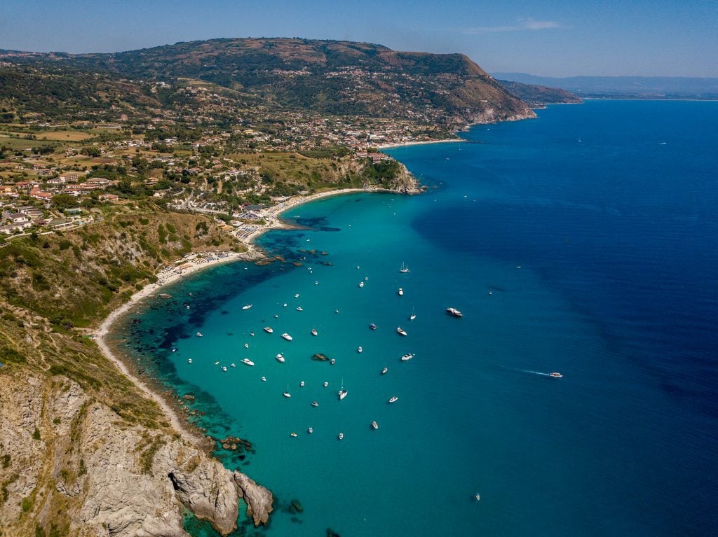 A beautiful coast in Italy ringed with golden beaches. In the water are about two dozen white boats.