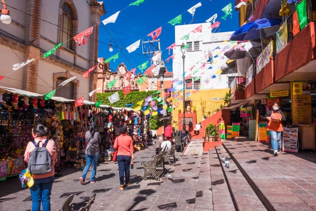 The outdoors next to a market with lots of stalls, people walking around, and red, white, and green paper flags hanging.