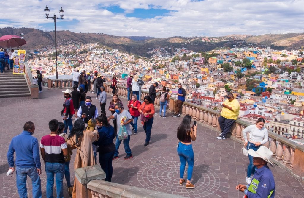 Around 30 people standing at the viewpoint overlooking the city, taking photos of each other.