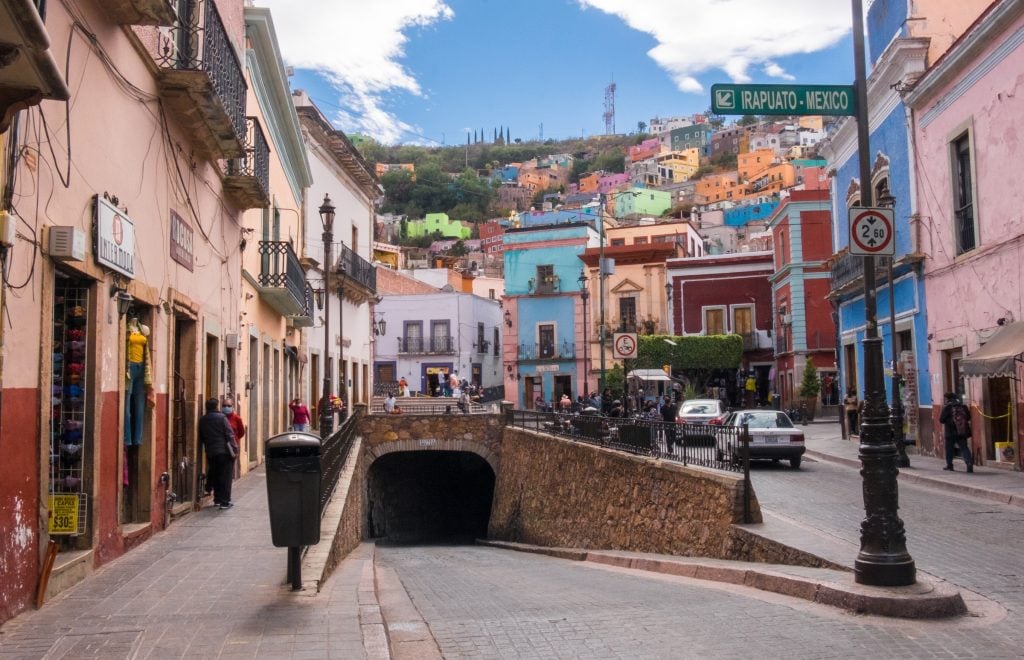 A small street square covered with colorful buildings, and in the foreground, a street leading into a stone tunnel.