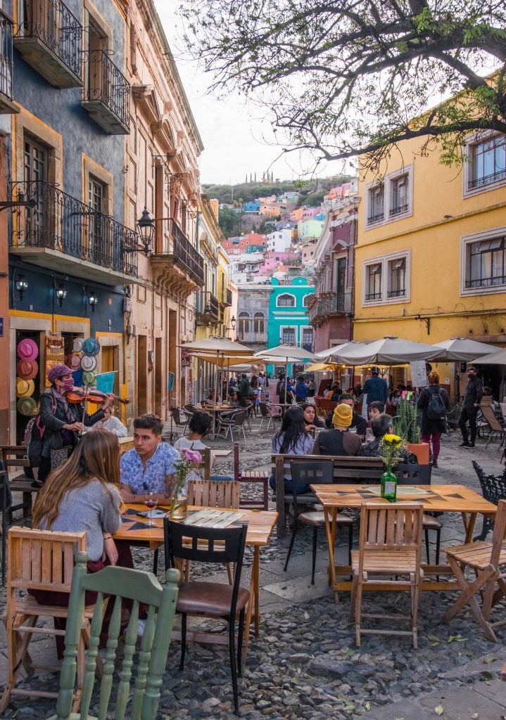 A small plaza with people eating outdoors, a violinist playing, and colorful buildings in the background.