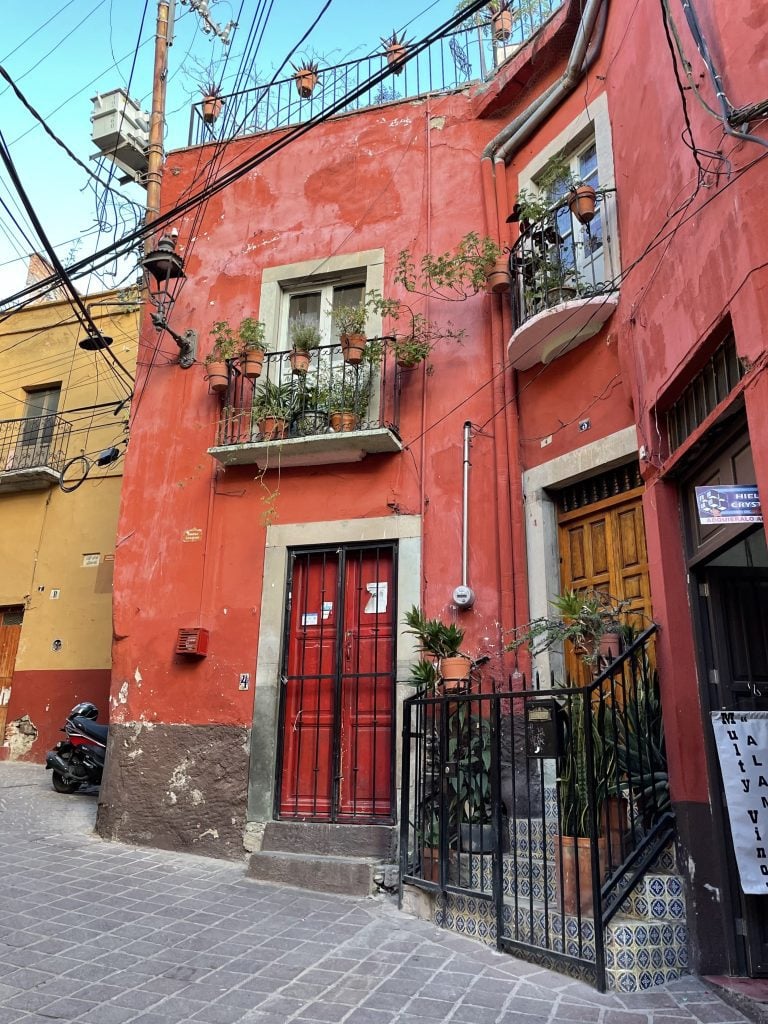 A pink house with a red door, and a white balcony above filled with lots of plants.