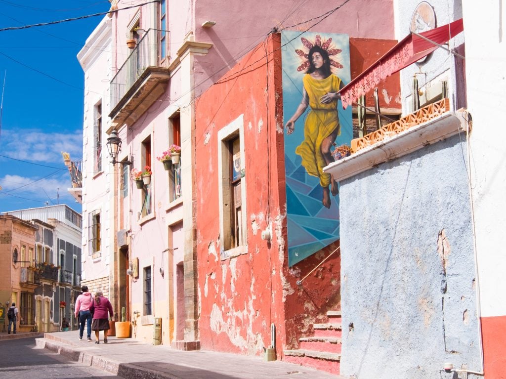 A colorful street with lots of pink buildings and a mural of an Indigenous woman in a yellow robe with a halo made of corn.