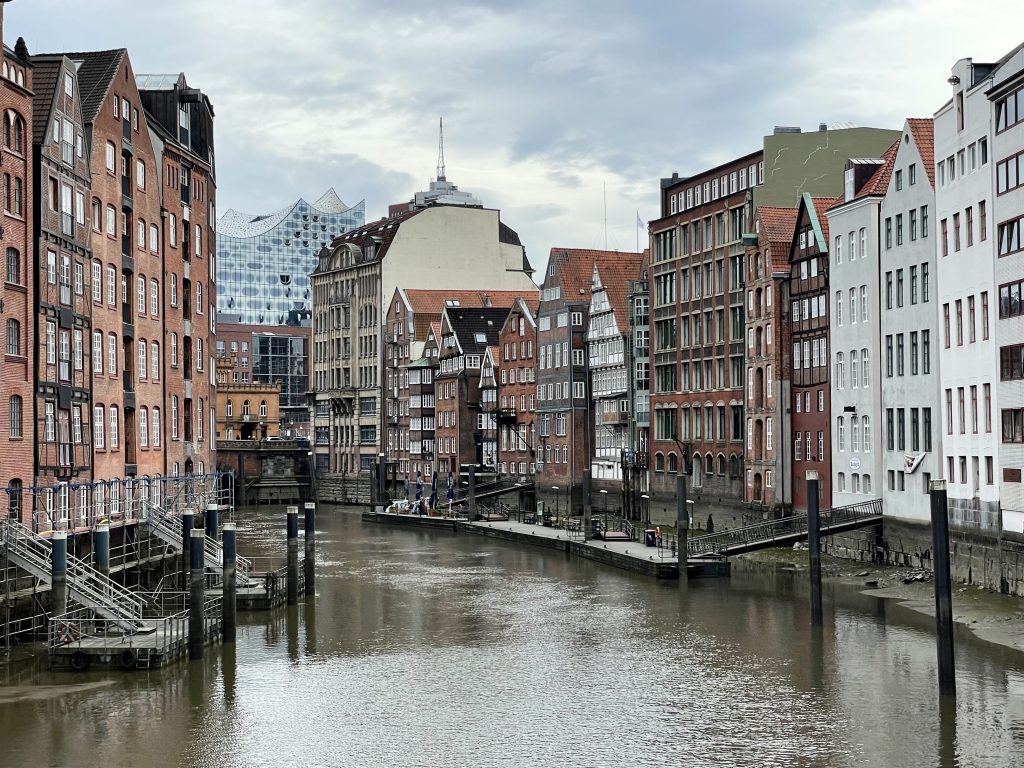 A canal in Hamburg surrounded by stately brown and white buildings with lots of windows. It's a cloudy day and the canal is brownish gray. In the background you see the blue-green fish scales of the philharmonic building.