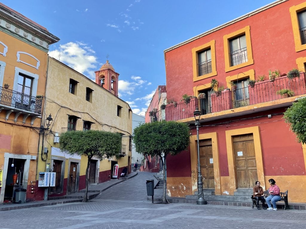 A small square edged with yellow and red buildings, two women sitting together on a bench.