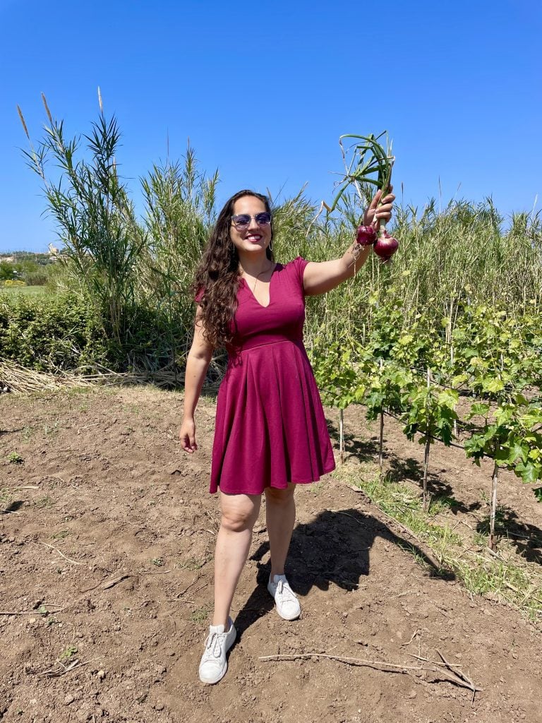 Kate stands in an onion field and holds up two purple onions, smiling big. She wears sunglasses, a burgundy short dress the same color as the onions, and white sneakers.