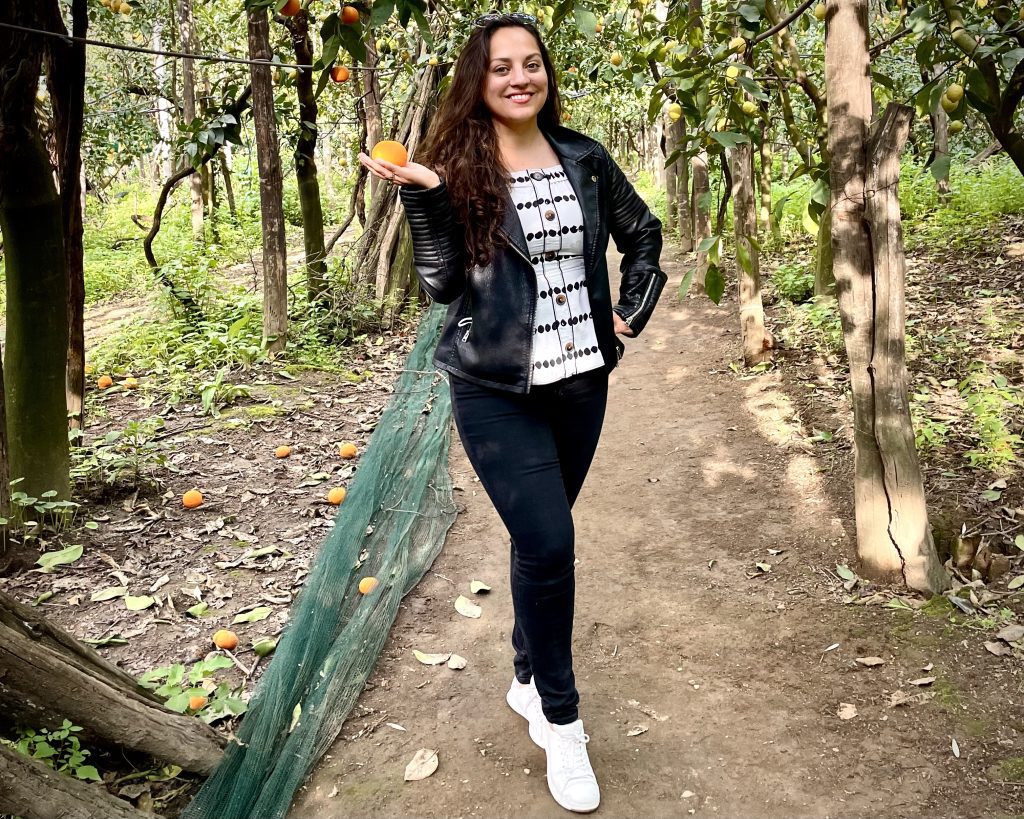 Kate standing in a lemon and orange grove, holding an orange in her hand. She wears a black leather jacket, white silk blouse with black polka dots, black jeans, and white sneakers.
