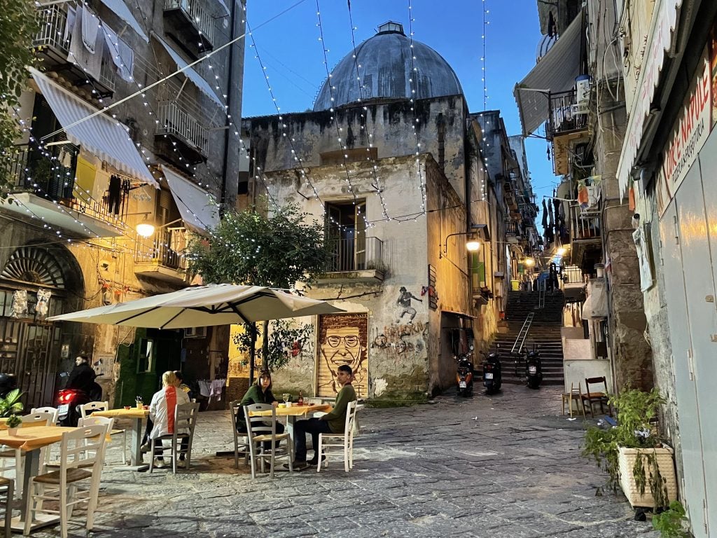 A small square in Naples just after sunset. People sitting at tables drinking wine, lights strung between buildings, laundry hanging off balconies, plants and graffiti everywhere.