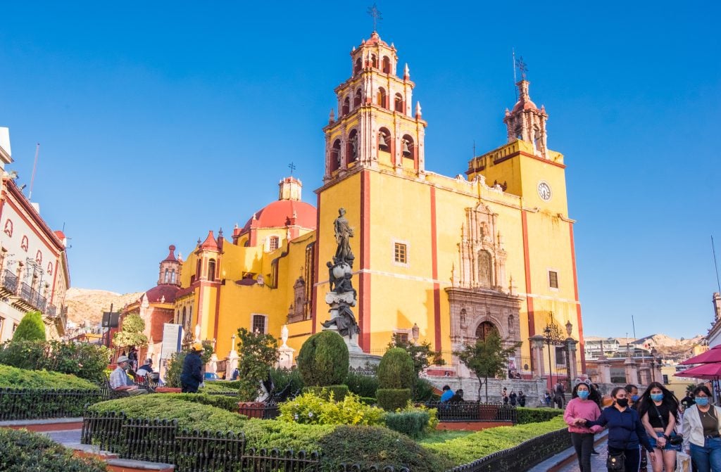 A bright yellow church with red trim, a small garden in front, and lots of people walking around with masks.