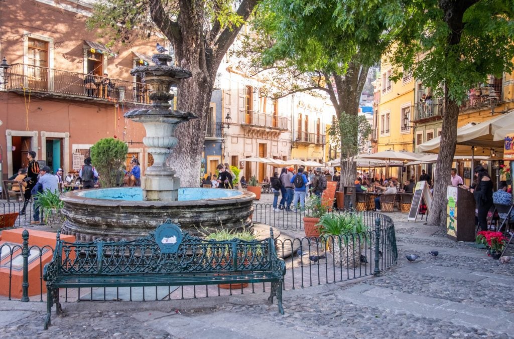 A large cobblestoned plaza with park benches, a fountain, and lots of outdoor cafes.