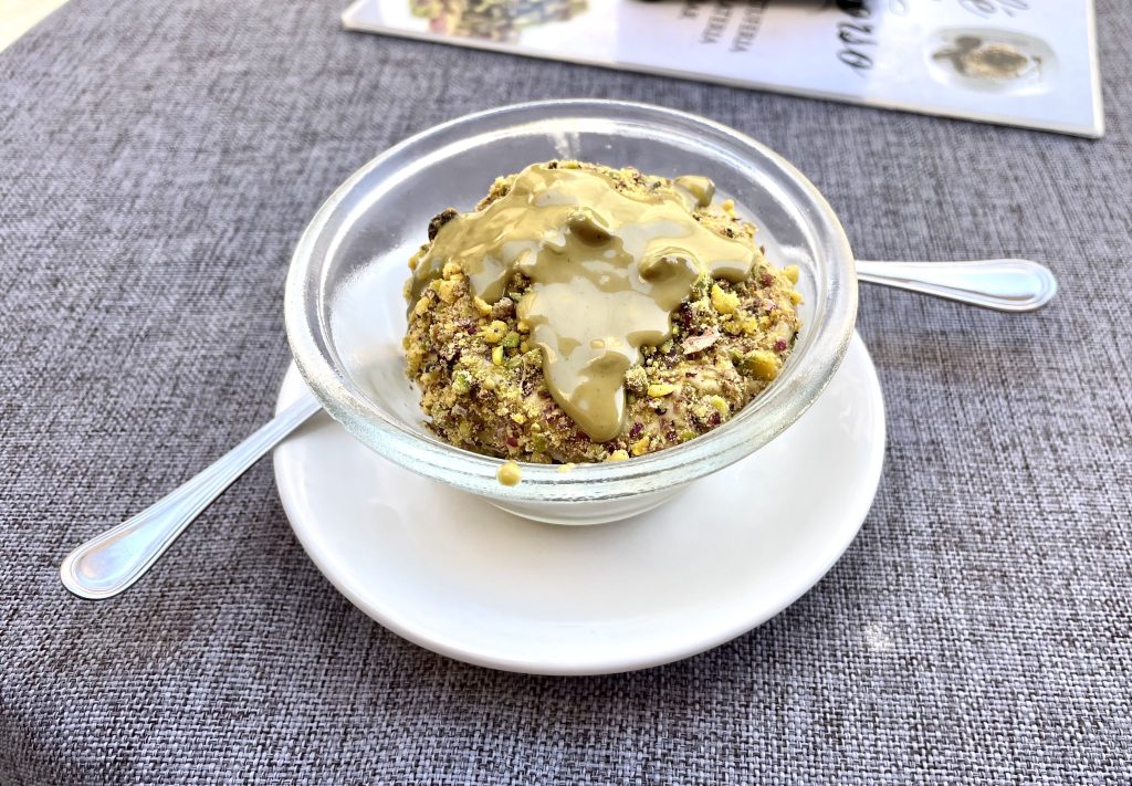 A bowl of pistachio ice cream topped with pistachio nuts and pistachio sauce, on a plate with two spoons.