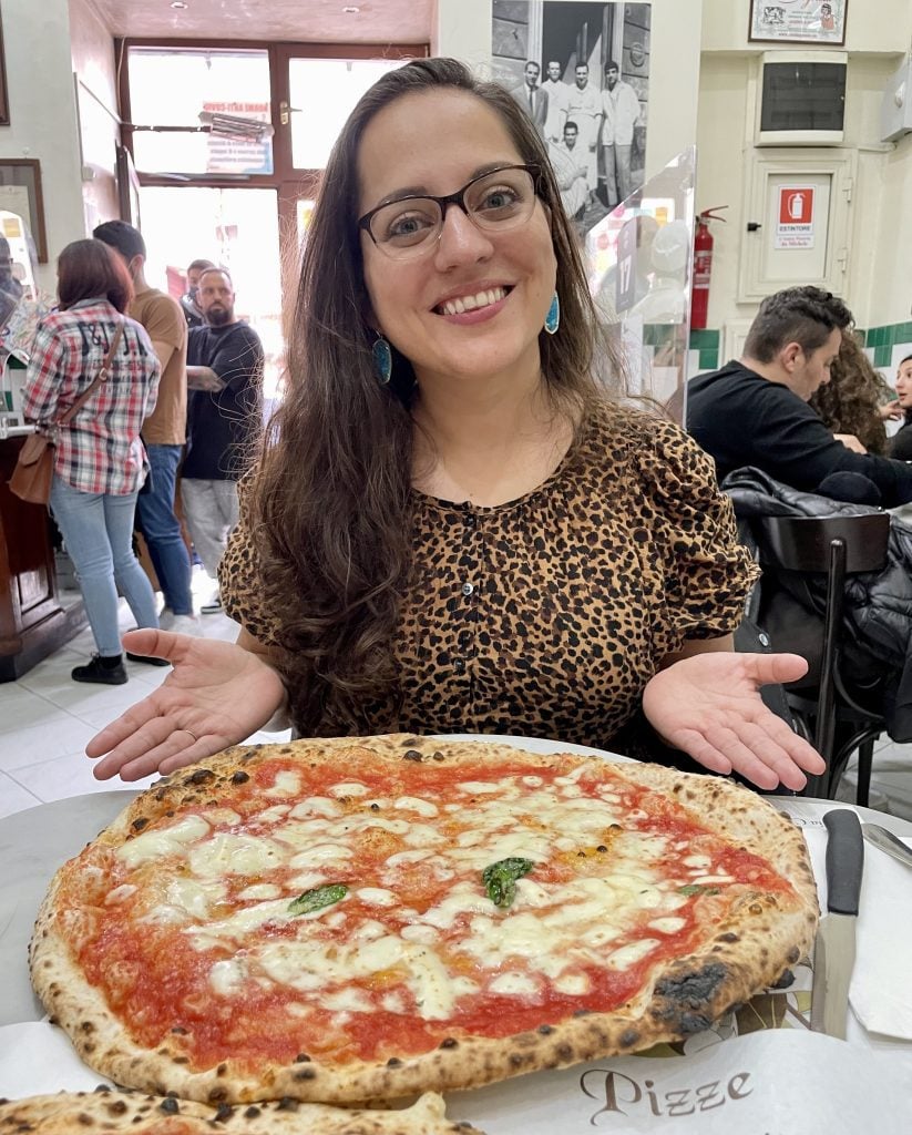 Kate smiling in front of a giant Margherita pizza. She wears glasses and a leopard-print shirt.
