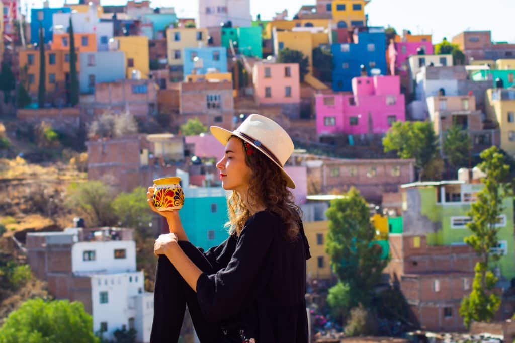 A woman in a Panama hat enjoying a cup of coffee, the colorful square homes of Guanajuato lit up behind her.