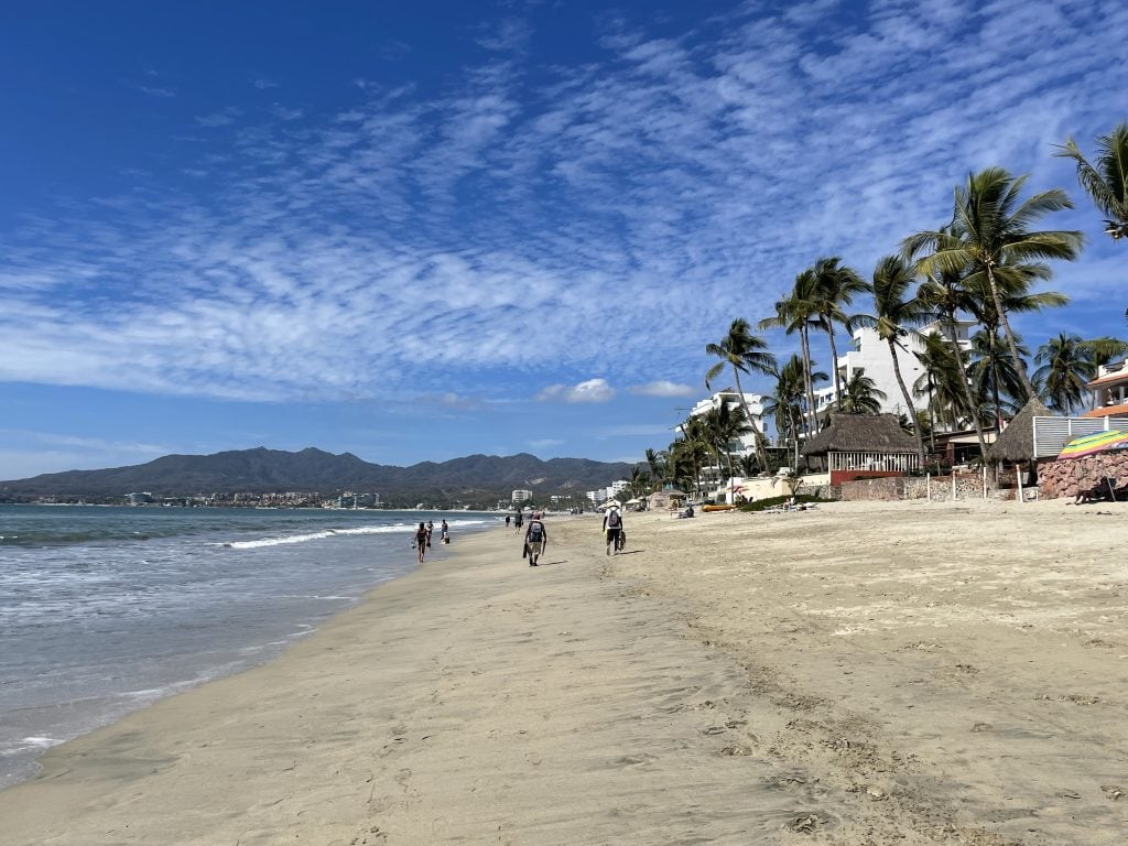 Beach vendors walking down a long expansive beach next to a placid ocean with small waves, mountains in the distance.