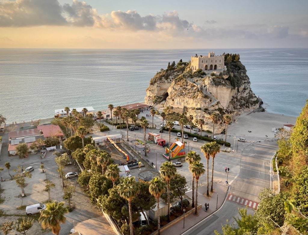 A large rocky outcropping close to the water, topped with a stone church, a long staircase leading up to it. The sun is about to set and the water looks warm and calm.