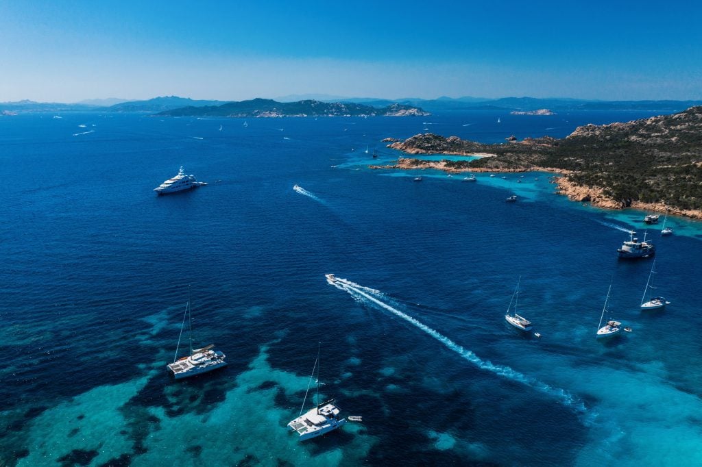 A blue sea scene in Sardinia with lots of white sailboats and a rocky coast. Looks like an archipelago.