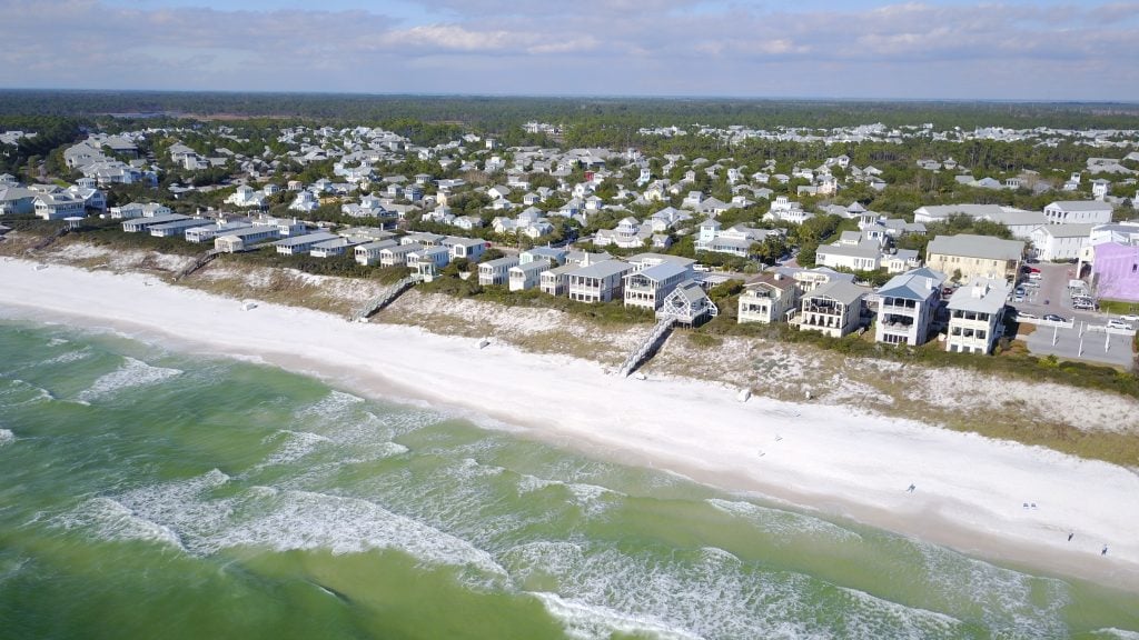 An aerial view of a long white beach with green ocean. Inland are small bungalows in a row.