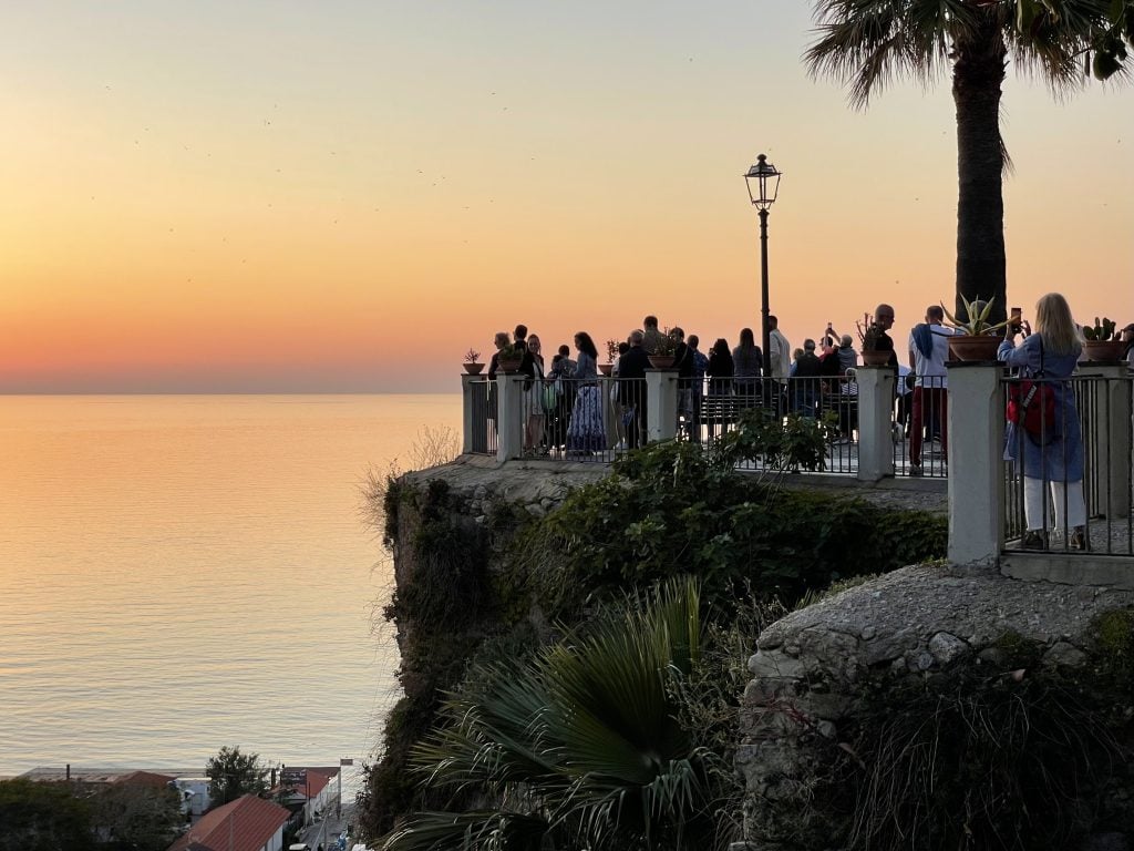 A group of people standing at a piazza atop a cliff, watching a purple, orange and yellow sunset over the sea.