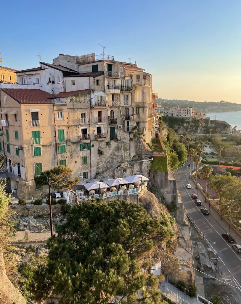 A view of a tall stone building built into a cliff, below it a restaurant with white umbrellas, and on the ground, a road running next to the ocean.