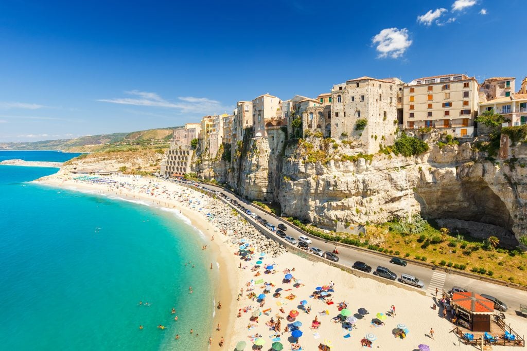 Clifftop stone buildings built into a cliff, overlooking a long sandy beach and bright turquoise water.